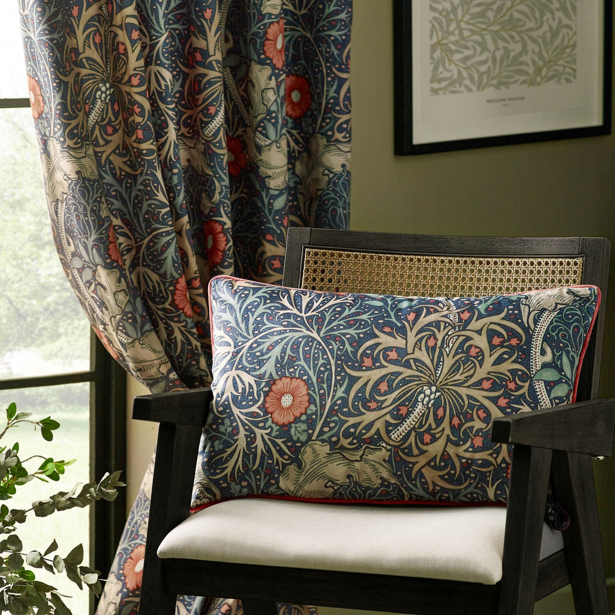 Seaweed fully lined velvet pencil pleat curtain in indigo with a Morris & Co. floral pattern, shown hanging by a window beside a matching printed cushion on a chair.