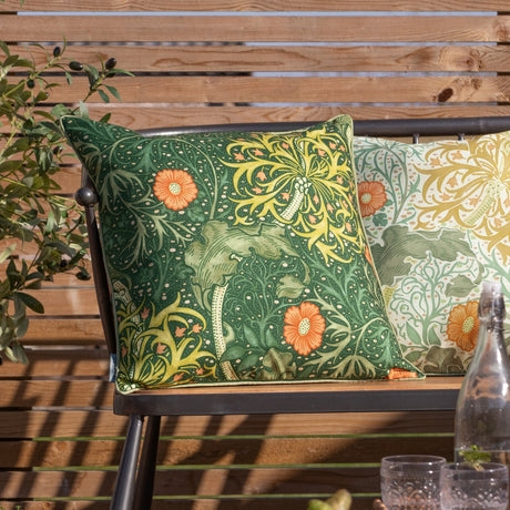 Square outdoor cushion in Morris & Co. Seaweed floral design, forest green fabric with sage and gold foliage and coral-orange blooms, shown on a garden bench.