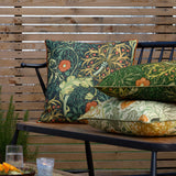 Seaweed Floral polyester cushion in forest green with sage and gold foliage and coral-orange blooms, shown among patterned cushions on an outdoor bench.