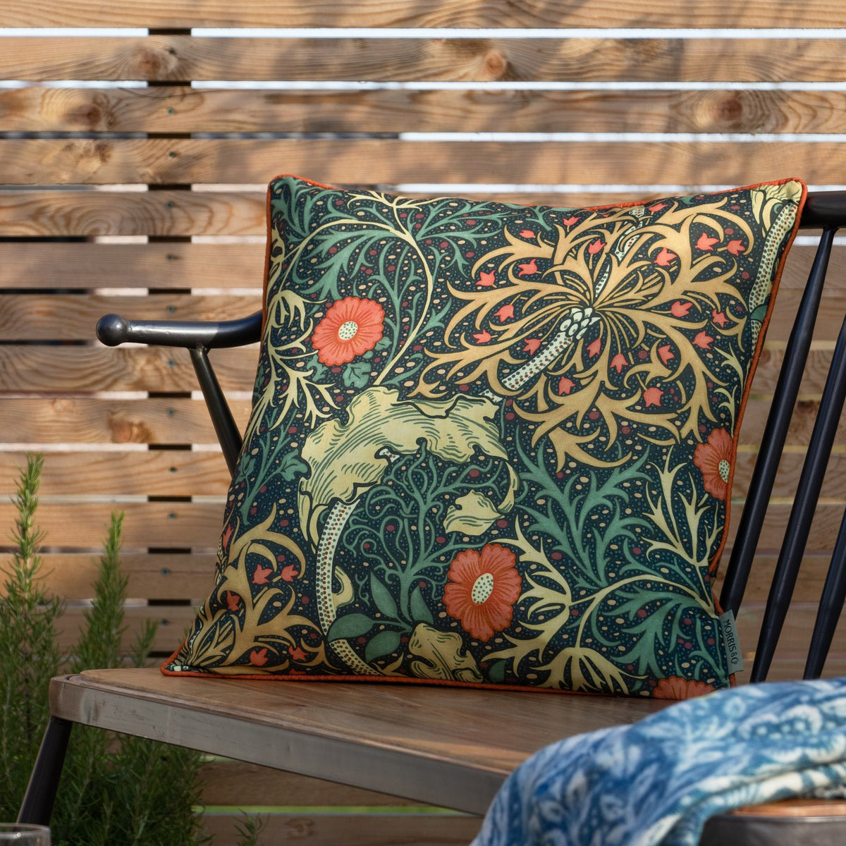 Square outdoor cushion in the Seaweed Floral pattern, indigo ground with orange daisy flowers and gold-green foliage, orange piped edge, shown on a garden bench.