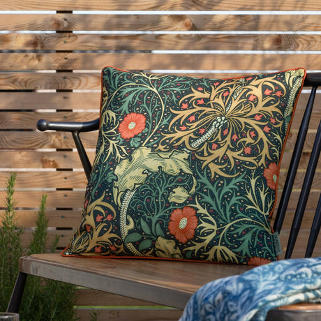Square outdoor cushion in the Seaweed Floral pattern, indigo ground with orange daisy flowers and gold-green foliage, orange piped edge, shown on a garden bench.