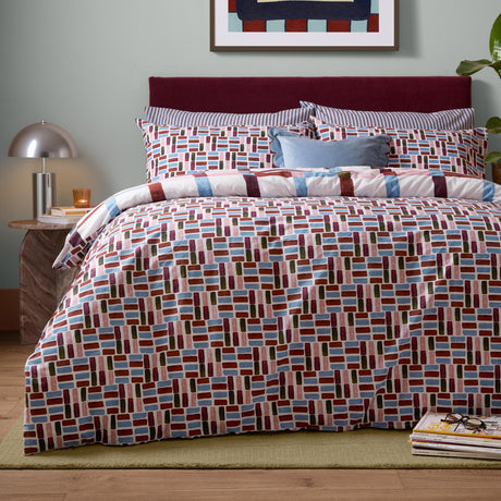 Selvan Reversible Check Duvet Cover Set in Oatmilk & Burgundy, geometric tile pattern in red, blue, olive and pink shown on a bed with striped pillowcases and a burgundy headboard.