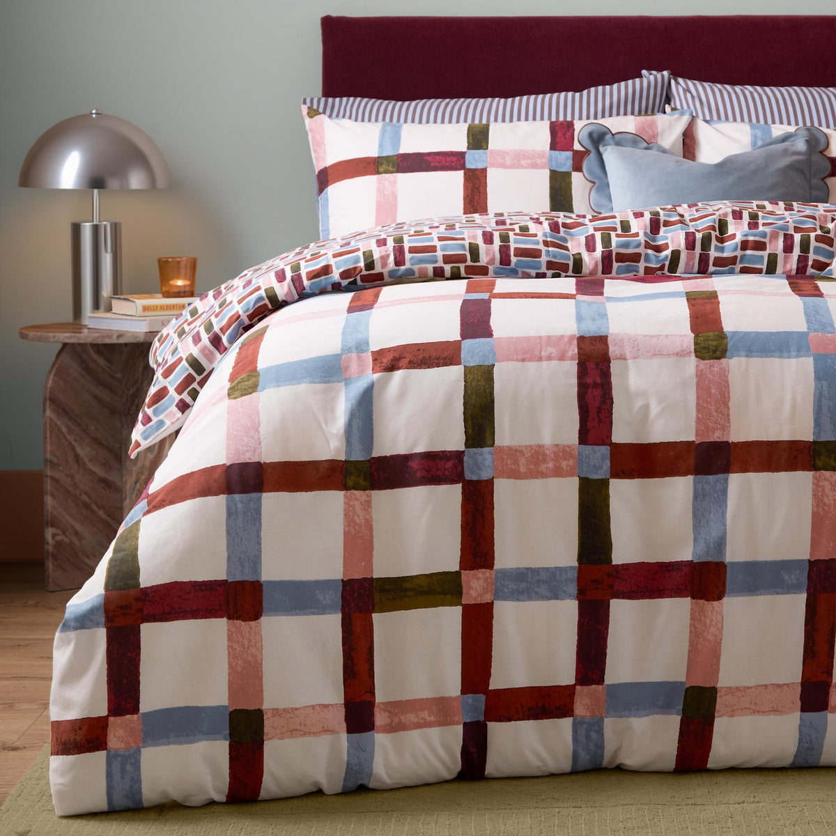 Selvan Reversible Check Duvet Cover Set in Oatmilk & Burgundy, featuring a painterly red, blue, olive and pink check, shown with striped and tiled-print pillowcases in a bedroom.