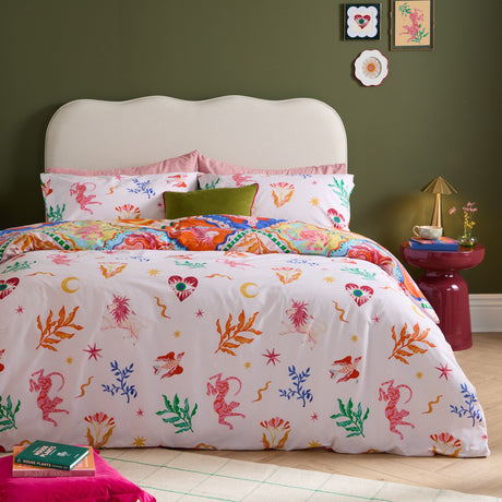 Seraphine Reversible Abstract Duvet Cover Set in Multi, shown on a bed with white side scattered with pink, orange and green mythical creatures and celestial motifs, styled in a green bedroom.