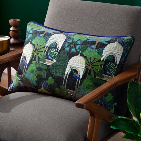 Serenity Velvet Cotton Cushion in Midnight, a rectangular cotton velvet cushion with peacocks and domed pavilions in green and blue on black, shown on a grey armchair.