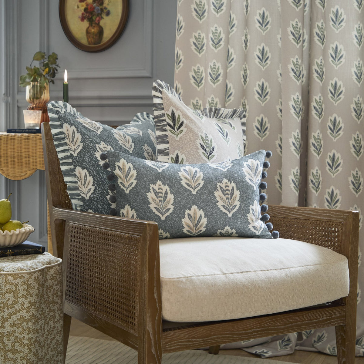Sessile Leaf Reversible Cotton Cushion in Delph Blue with cream leaf motif and pom-pom trim, styled on a cane armchair among matching floral cushions and curtain.