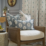 Sessile Leaf Reversible Cotton Cushion in Delph Blue with cream leaf motif and pom-pom trim, styled on a cane armchair among matching floral cushions and curtain.