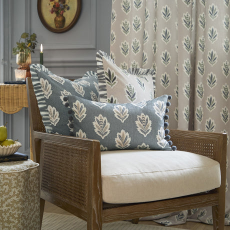 Sessile Leaf Reversible Cotton Cushion in Delph Blue with cream leaf motif and pom-pom trim, styled on a cane armchair among matching floral cushions and curtain.