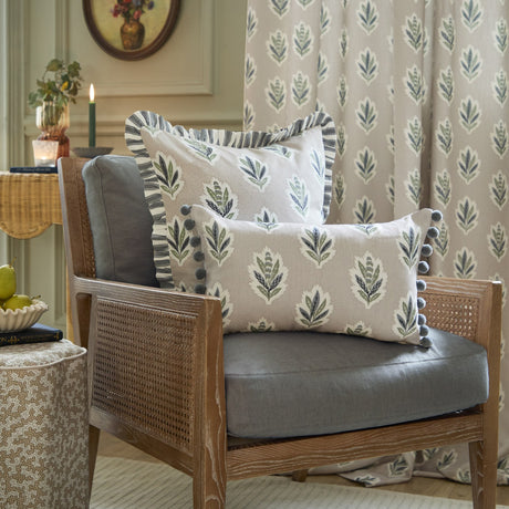 Sessile Leaf reversible cotton cushion in flax with green and slate blue leaf motif, shown on a rattan and grey upholstered armchair with a matching lumbar cushion.