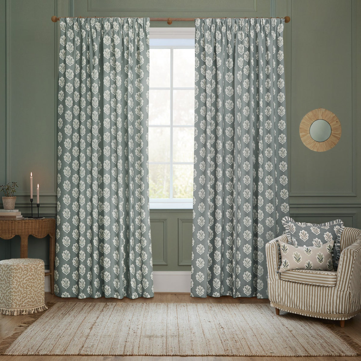 Sessile Leaf reversible cotton cushion in flax, shown on a striped armchair in a living room styled with matching green leaf-print curtains.
