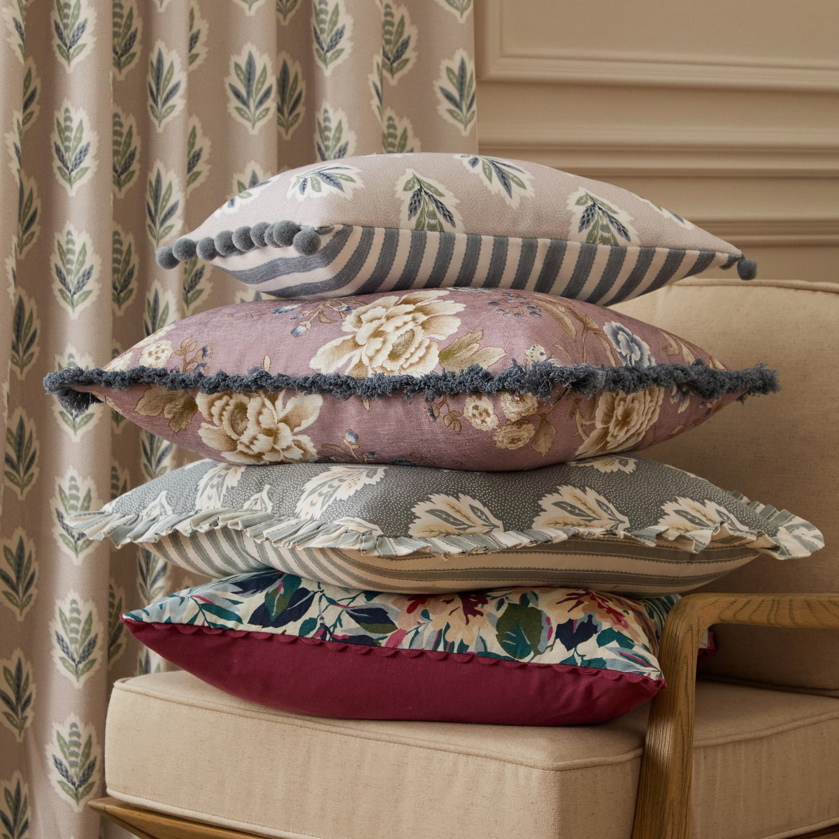 Sessile Leaf Reversible Cotton Cushion in Flax with pompom trim, shown top of a stacked pile of patterned cushions on a chair beside leaf-print curtains.