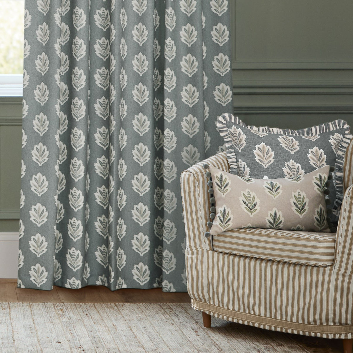 Sessile Leaf pencil pleat curtains in Delph Blue with a stacked oak leaf motif, shown as pencil-pleat drapes beside a striped armchair with matching leaf-print cushions.
