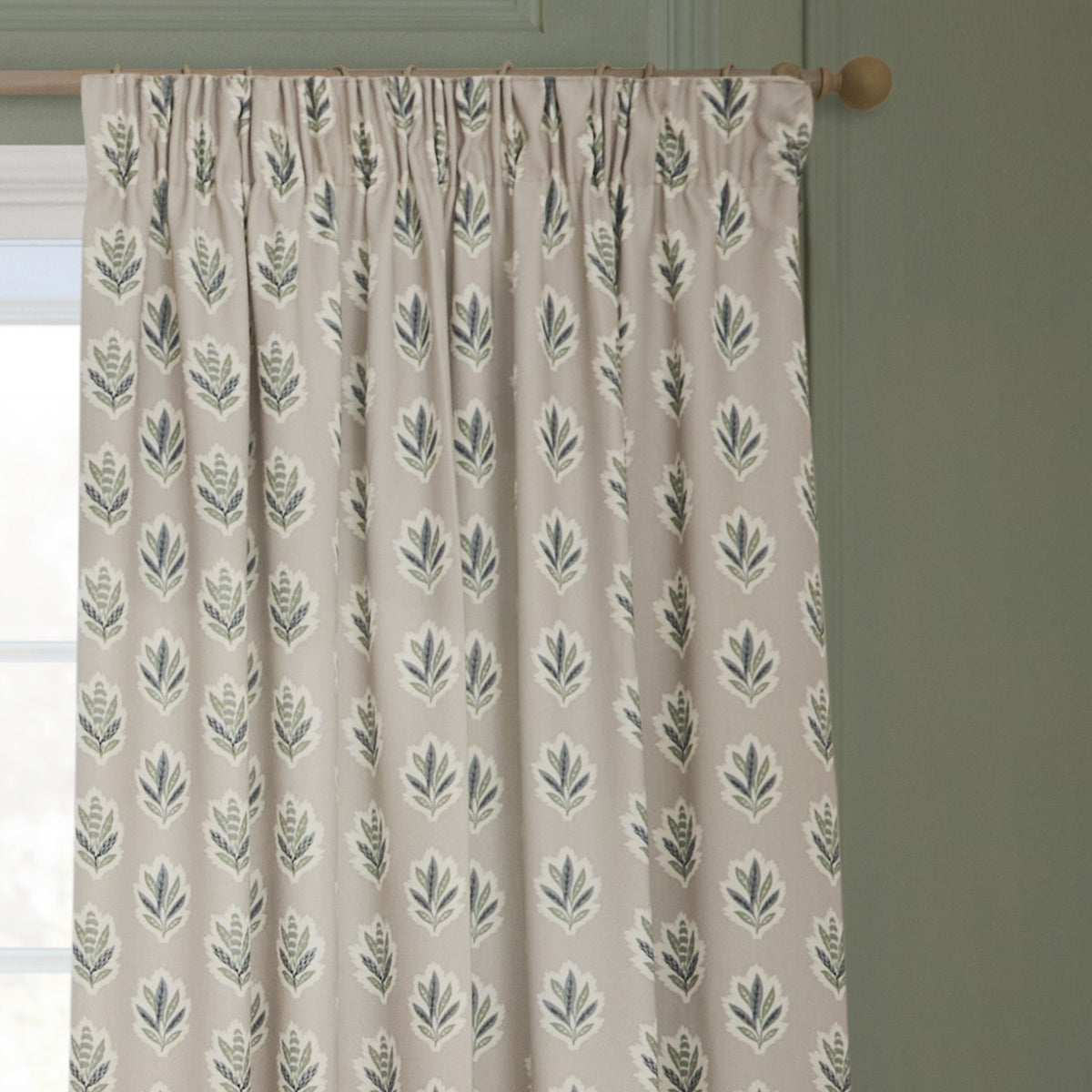 Sessile Leaf pencil pleat curtains in flax beige polyester with a green and slate blue oak-leaf print, shown gathered on a wooden pole against a green wall.