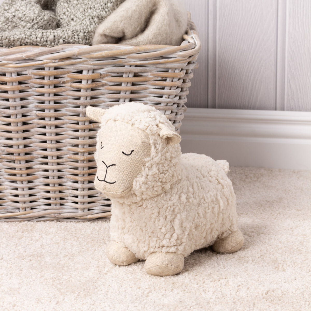 Sheep Wool Cotton Door Stop in white, with curly sherpa body and woven herringbone face, ears and feet, shown on cream carpet beside a wicker basket.