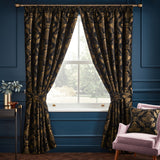Shiraz pencil pleat curtains in black with gold jacquard floral damask, shown as a tied-back pair with matching cushion in a navy-panelled living room.