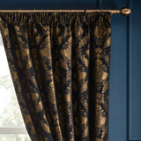 Shiraz pencil pleat curtains in black chenille jacquard fabric with a gold floral damask pattern, shown hanging on a bronze curtain pole against a blue wall.