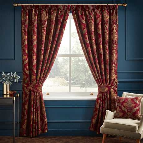 Shiraz Jacquard Chenille pencil pleat curtains in burgundy with a gold and green floral damask pattern, shown with matching tiebacks and cushion in a dark blue living room by a window.