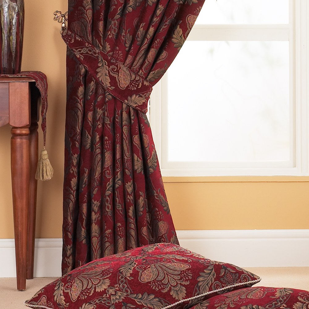 Pencil pleat curtain in burgundy jacquard chenille with a gold and green floral damask pattern, shown tied back beside a window, with matching cushions in the foreground.