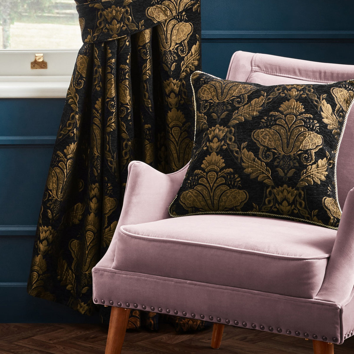 Shiraz Jacquard Chenille Cushion in black with gold floral damask pattern and corded edge, shown on a pink velvet armchair beside matching curtains.