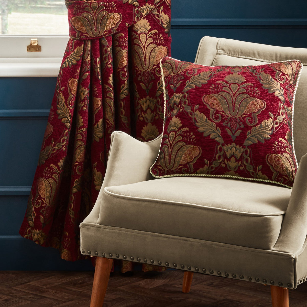 Shiraz Jacquard Chenille Cushion in burgundy with gold and olive damask floral pattern, shown on a beige velvet armchair beside a matching curtain.
