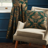 Shiraz Jacquard Chenille Cushion in emerald green with a gold floral damask pattern and rope-trimmed edge, shown on a beige armchair beside matching curtains.