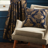 Shiraz Jacquard Chenille Cushion in navy with gold floral damask pattern, shown on a beige armchair beside matching patterned curtains in a living room setting.