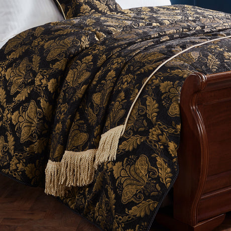 Shiraz Jacquard Chenille Throw in black with woven gold floral damask pattern and tasselled fringe, draped over a bed beside a dark wood bed frame.