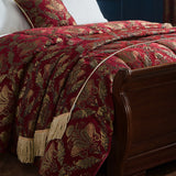 Shiraz Jacquard Chenille Throw in burgundy red with gold floral damask pattern and twisted gold fringe edging, draped over a bed with matching cushion shams.