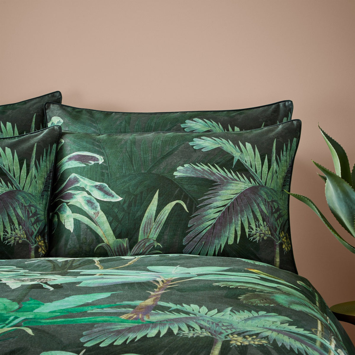 Siona Cotton Jungle Duvet Cover Set in multi, shown with matching pillowcases in a dark green tropical palm print against a black ground, styled on a bed with a soft pink wall backdrop.