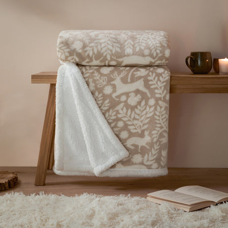 Skandi Woodland Printed Polyester Throw in Sable, folded on a wooden bench showing cream deer, hare and foliage patterns with a white sherpa fleece reverse, styled with a mug and candle.