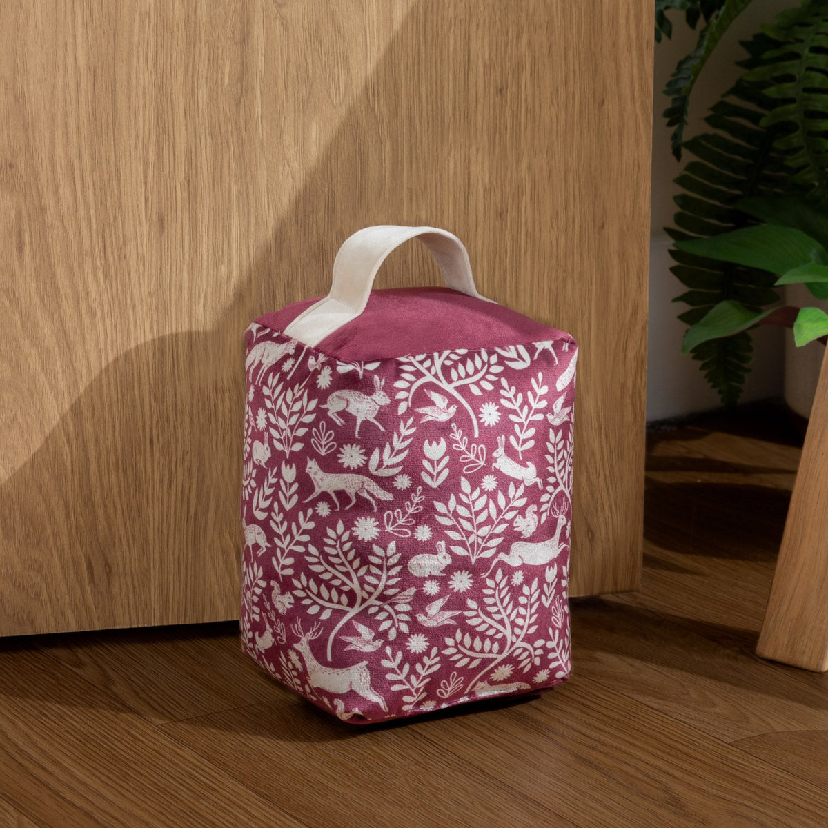 Skandi Woodland Animal Velvet Door Stop in berry, cream fabric handle, printed with hares, foxes and birds among leaves, shown against a wooden door and floor.