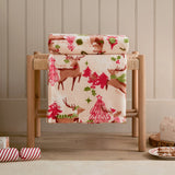Sleigh Team Printed Polyester Throw in cream draped over a wooden stool, showing a festive pattern of reindeer, pink Christmas trees and snowflakes in red, pink and green.