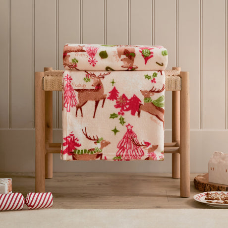 Sleigh Team Printed Polyester Throw in cream draped over a wooden stool, showing a festive pattern of reindeer, pink Christmas trees and snowflakes in red, pink and green.