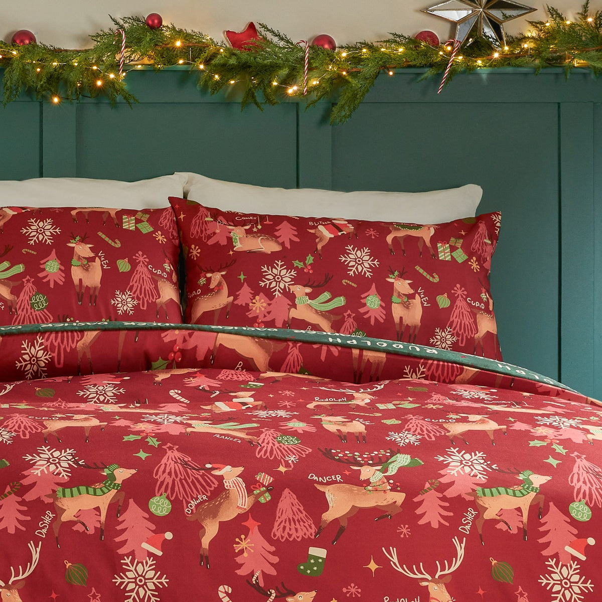 Sleigh Team Printed Reversible Duvet Cover Set in red, shown on a bedroom bed with matching pillowcases featuring scarved reindeer, Christmas trees and snowflakes among festive garland decor.