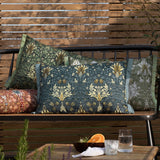 Snakeshead Floral polyester cushion in berry red with mirrored tan, cream and teal floral pattern, sage-green Oxford trim, shown on an outdoor bench among other patterned cushions.