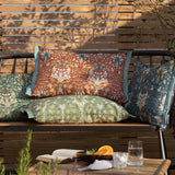 Snakeshead Floral polyester lumbar cushion in indigo with leaf-green foliage and cream-gold blooms, shown on a wooden garden bench with matching red and green patterned cushions.