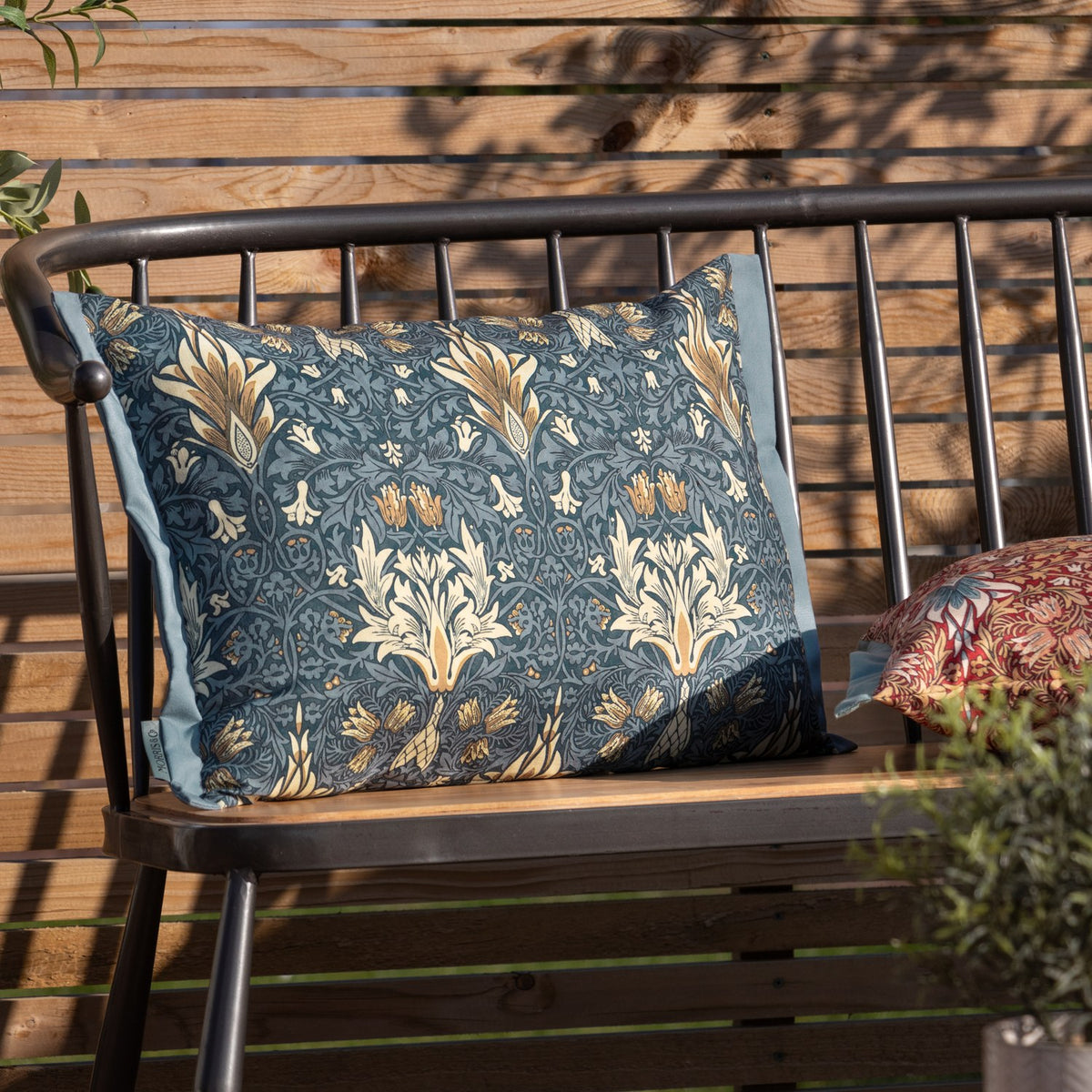 Rectangular Snakeshead Floral polyester cushion in indigo with cream and gold floral print, shown on a metal-framed bench with wooden slats in an outdoor setting.