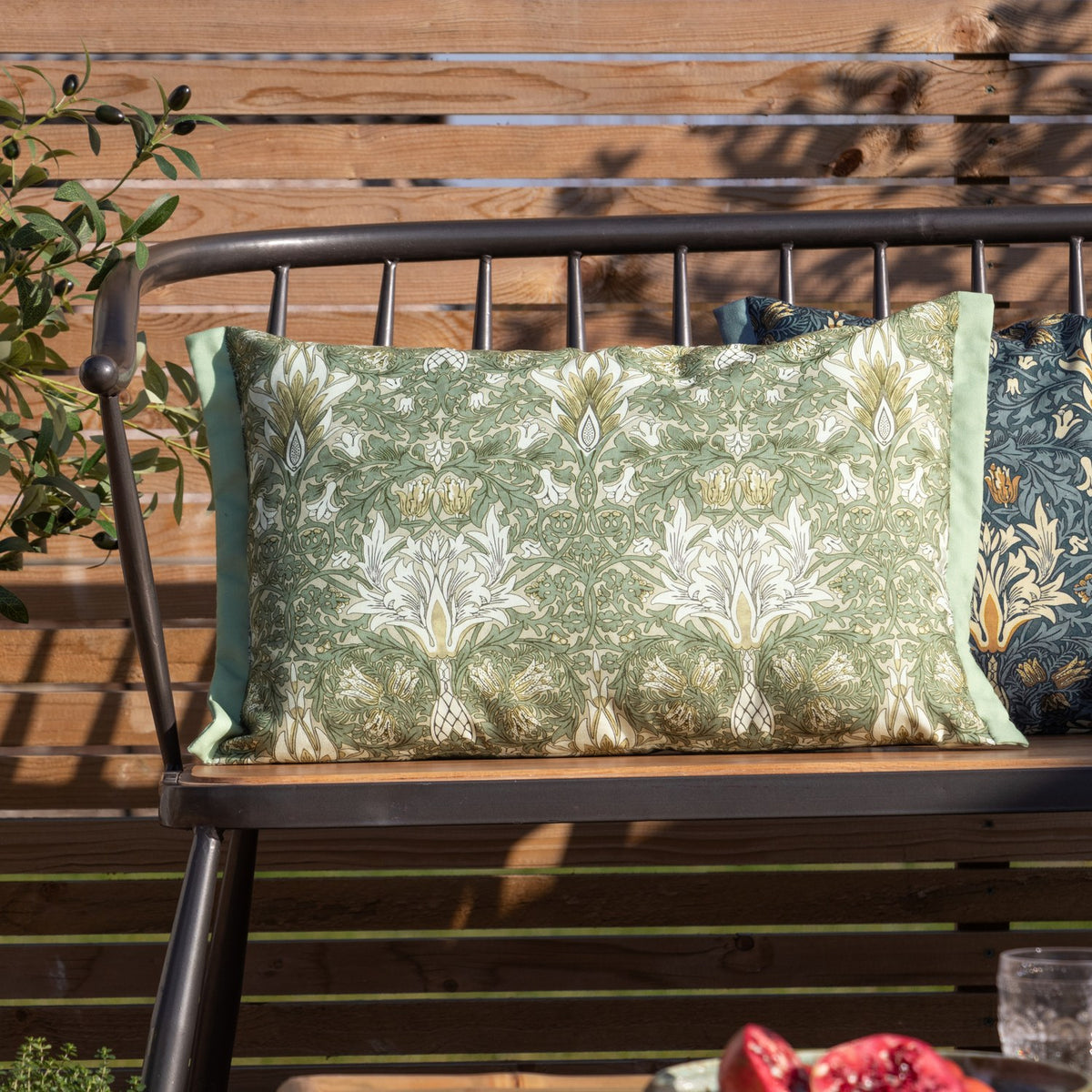 Rectangular Snakeshead Floral polyester cushion in green, gold and cream on a stone ground, with mint piped edges, shown on an outdoor bench.