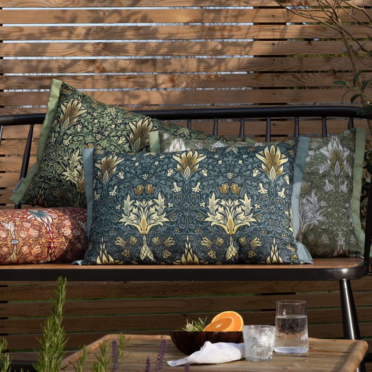 Rectangular Snakeshead Floral polyester cushion in gold, green and cream on a stone ground, shown among patterned pillows on an outdoor bench.