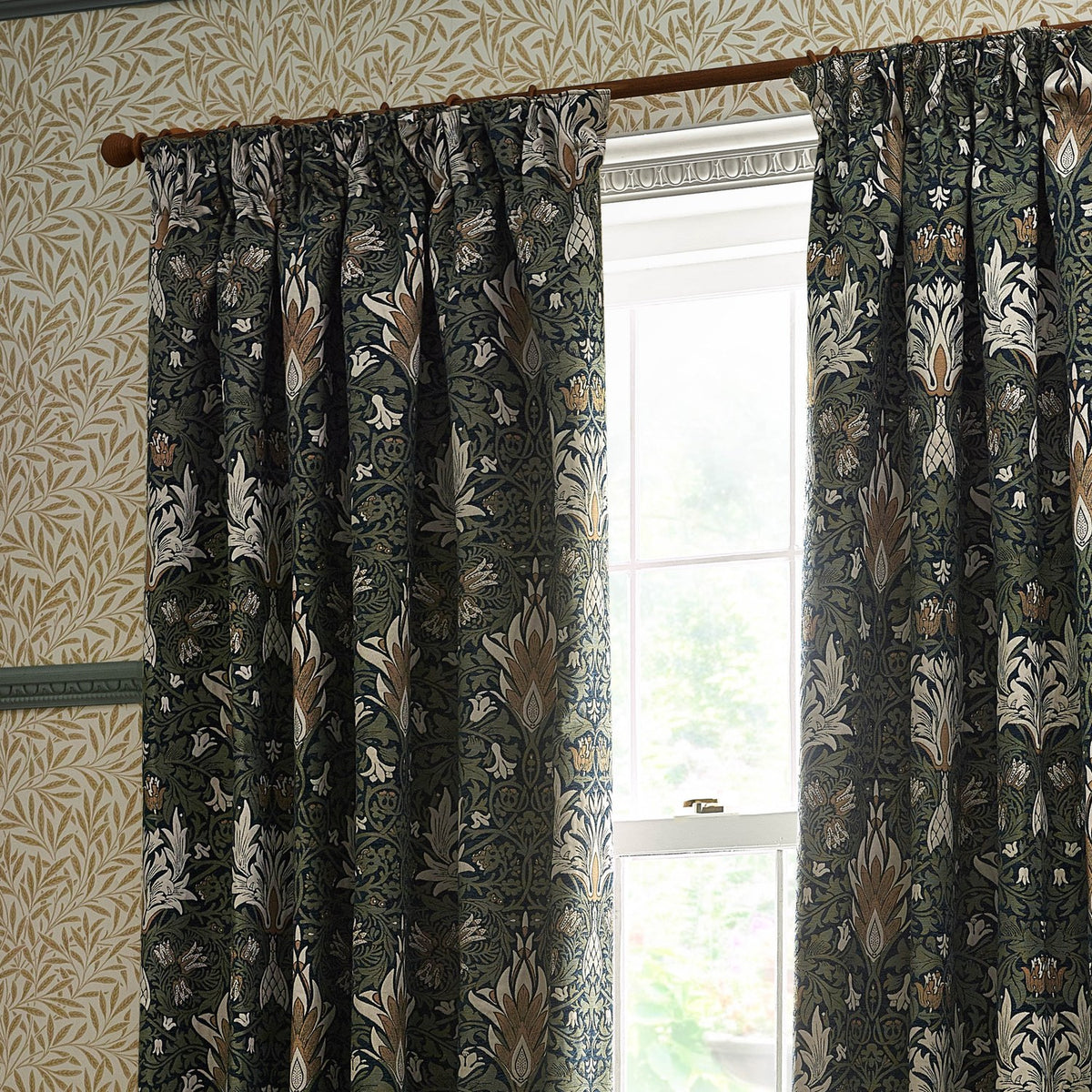 Snakeshead Jacquard Chenille pencil pleat curtains in Indigo/Leaf, floral fritillary pattern in navy, green and cream, shown pleated on a wooden pole beside a window.