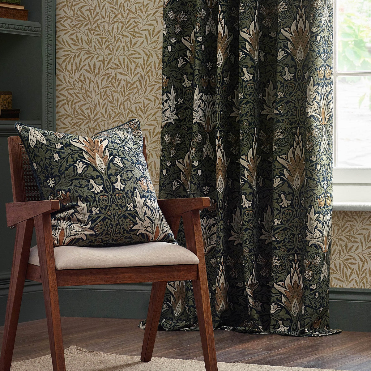 Snakes Head Jacquard Chenille Cushion in indigo and leaf green, styled on a wooden armchair against patterned wallpaper and matching curtains in a living room setting.