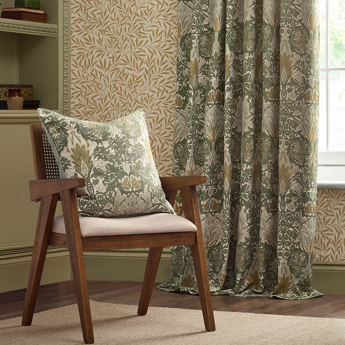 Snakes Head Jacquard Chenille Cushion in Sunflower/Stone on a wooden armchair, showing a sage green ground with cream foliage and gold flowerhead motifs, set beside matching curtains in a living room.