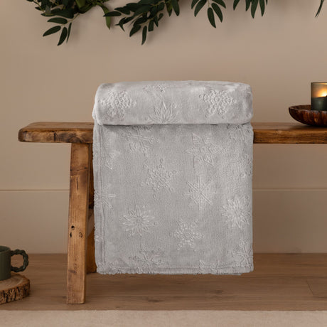 Grey faux fur throw with embossed snowflake pattern, folded over a wooden bench in a living room setting with candle and greenery nearby.