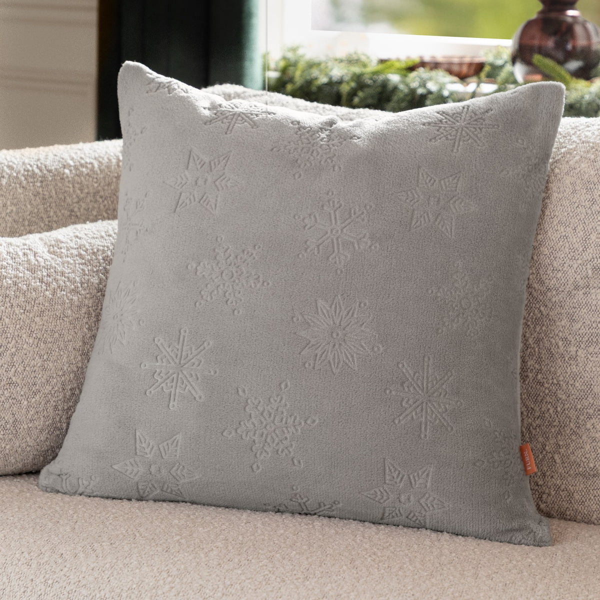 Square grey faux fur cushion with raised snowflake and star motifs, shown propped on a beige sofa in a living room setting.