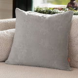 Square grey faux fur cushion with raised snowflake and star motifs, shown propped on a beige sofa in a living room setting.