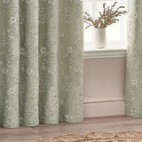 Sophia Jacquard Embroidered Eyelet Curtains in sage green with botanical floral pattern, shown framing a window beside a white vase in a living room setting.