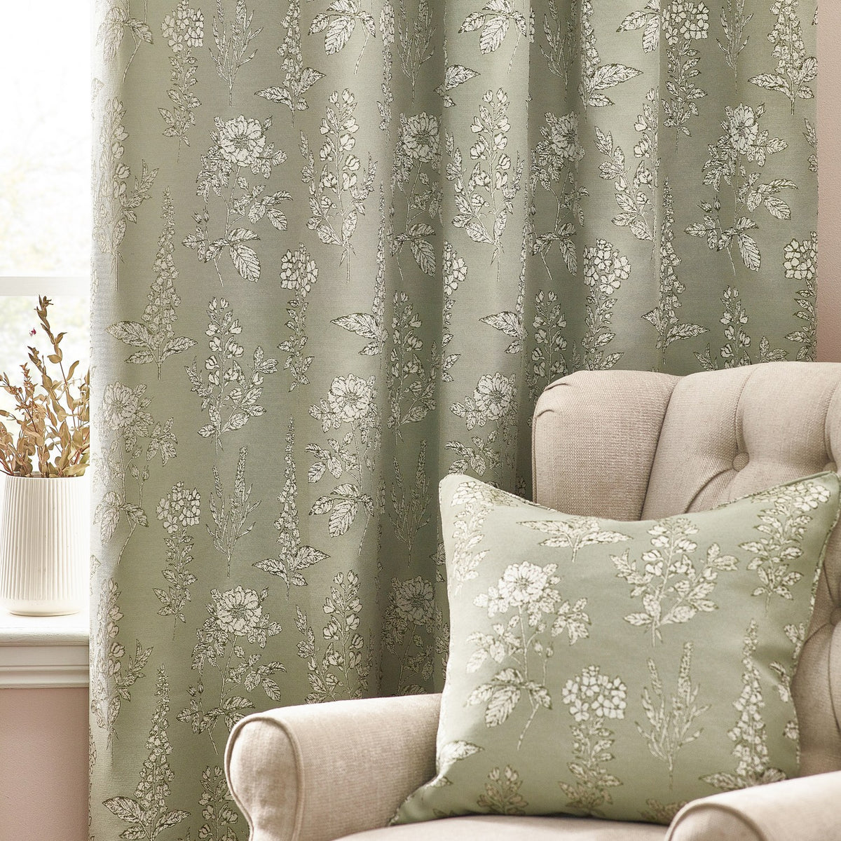 Sophia Jacquard Embroidered Eyelet Curtains in sage green with botanical foxglove and rose pattern, shown in a living room beside a matching cushion on a beige armchair.