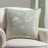 Sophia Eyelet Polyester Cushion in Sage, a 50 x 50cm square jacquard cushion with woven botanical florals and leaves in cream and olive, shown on a cream armchair.