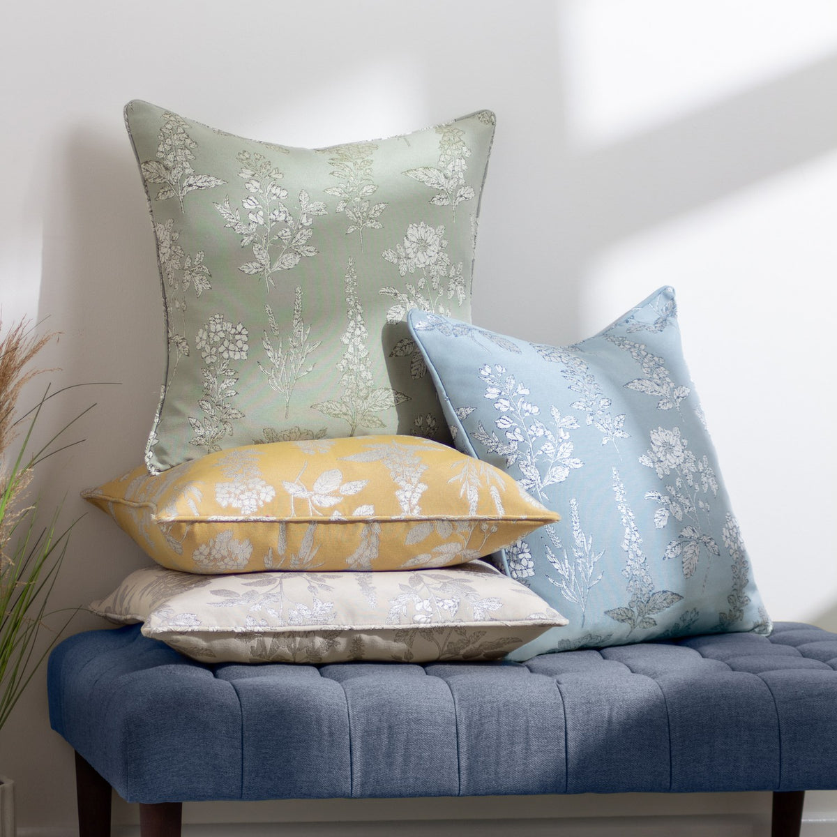 Sophia Eyelet Polyester Cushion in sage with woven botanical floral pattern, shown alongside blue, yellow and beige patterned cushions on a blue tufted bench.
