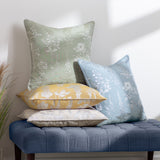 Sophia Eyelet Polyester Cushion in sage with woven botanical floral pattern, shown alongside blue, yellow and beige patterned cushions on a blue tufted bench.